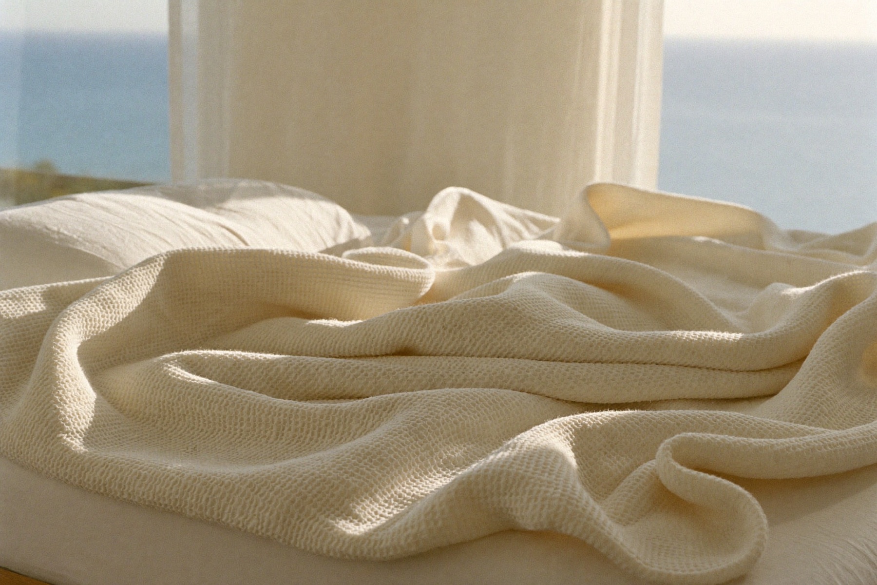 Oat-cream waffle muslin blanket draped across a white linen bed with an ocean view through the window